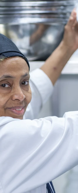 A female culinary student in a black headscarf and white uniform reaches for a stainless-steel mixing bowl on a kitchen shelf, smiling at the camera. A female culinary student in a black headscarf and white uniform reaches for a stainless-steel mixing bowl on a kitchen shelf, smiling at the camera.